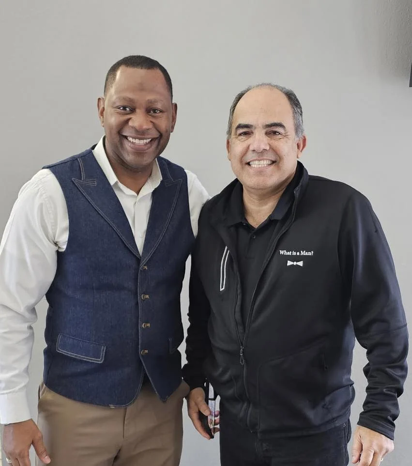 Two men smiling at camera against white background, one wearing denim vest and white shirt, other in black jacket