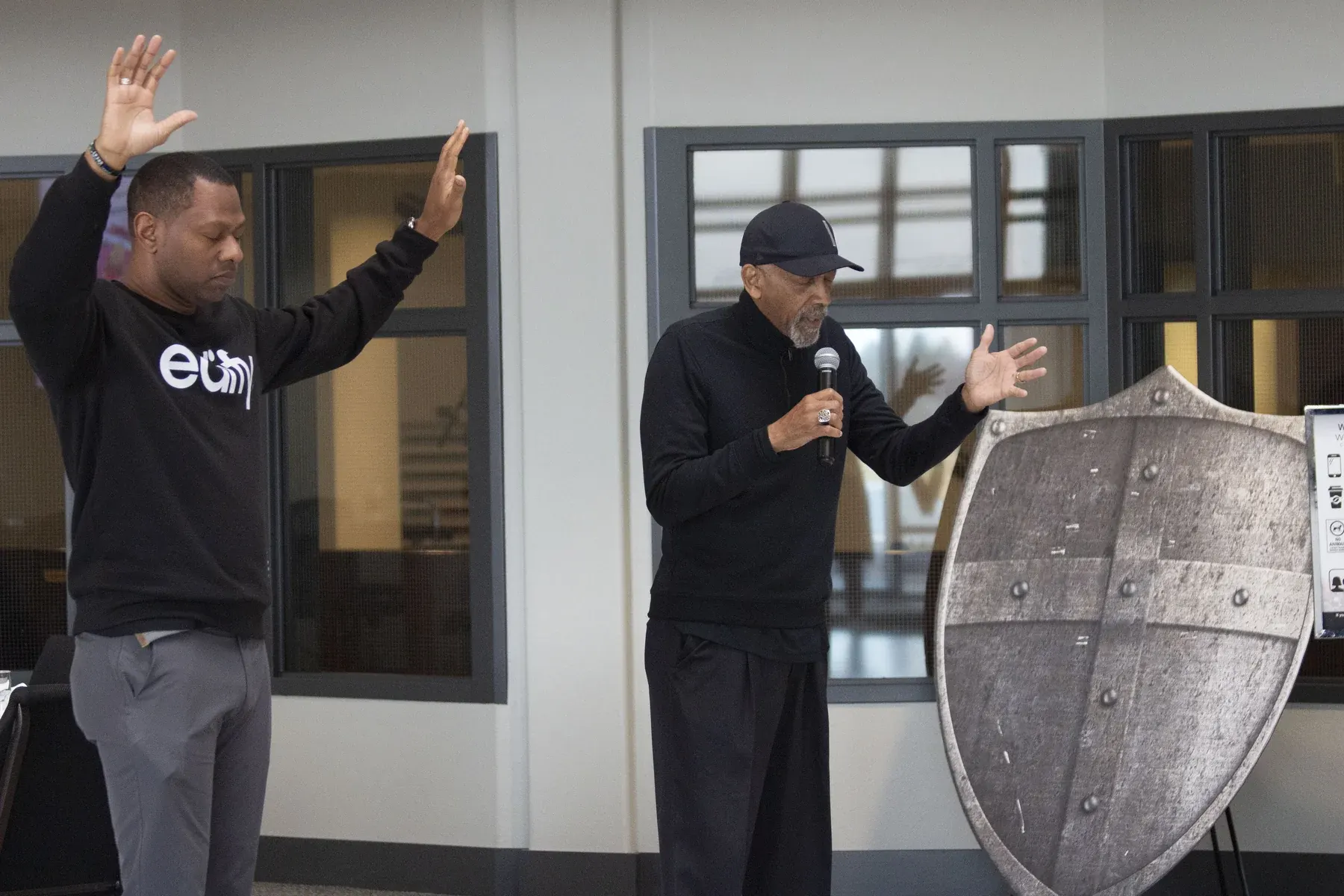 Two men speaking at an indoor event, one holding a microphone near a large wooden shield display
