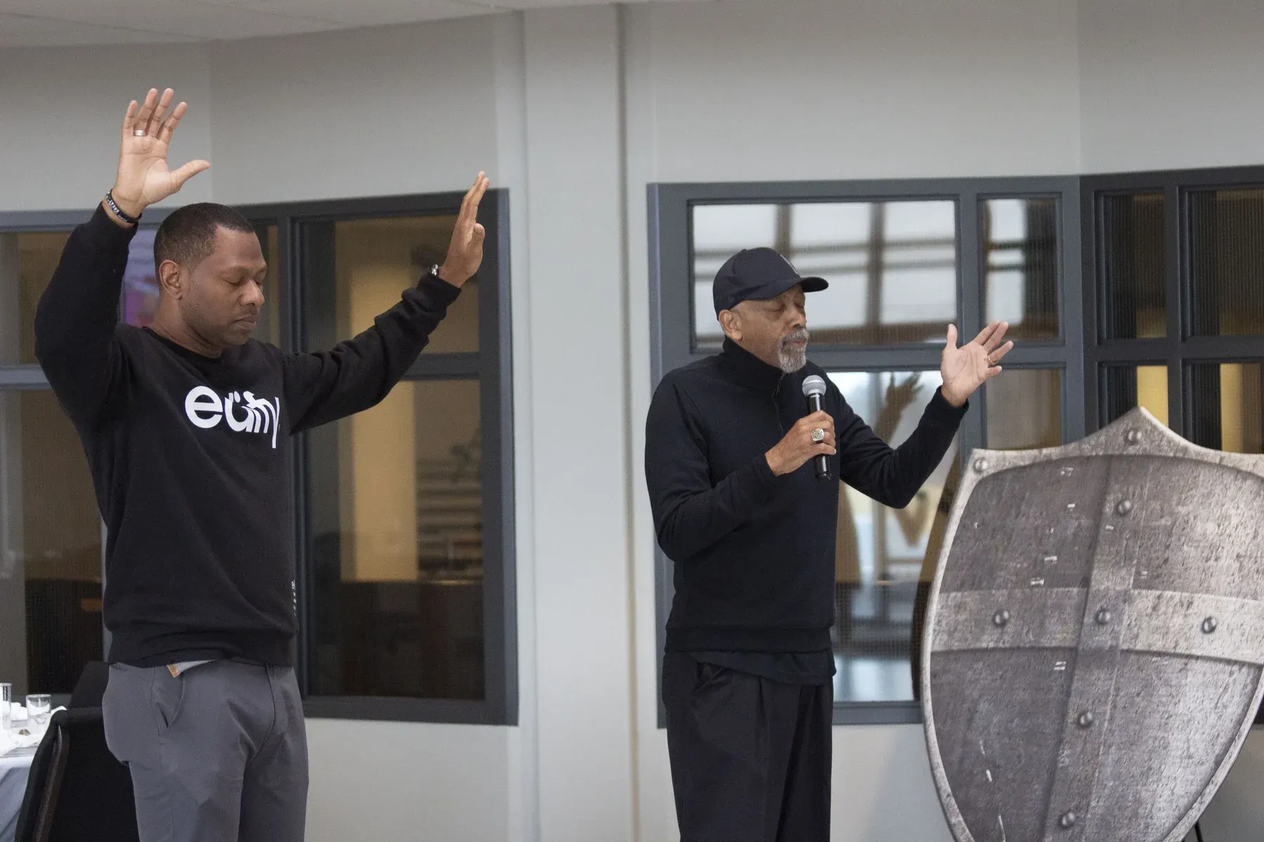 Two men speaking at indoor event, one holding microphone with hands raised
