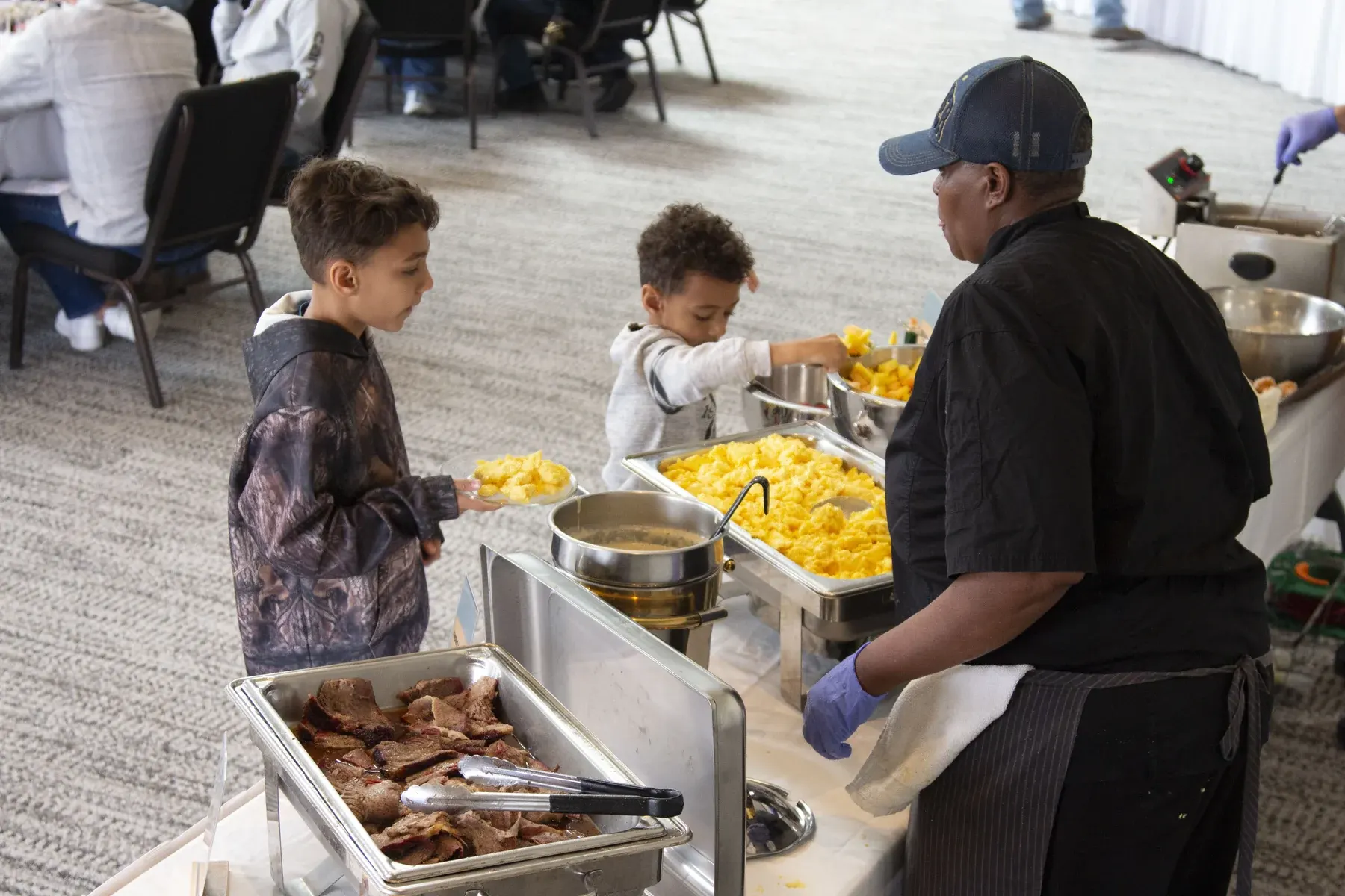 Man in black shirt serving corn to two young children at outdoor food buffet