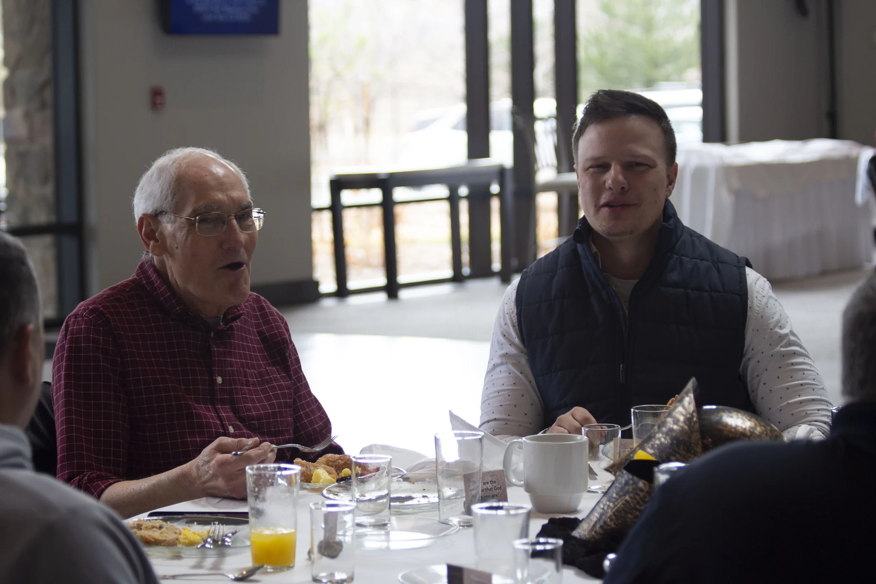 Older man in maroon shirt and younger man in navy vest seated at dining table during business meeting