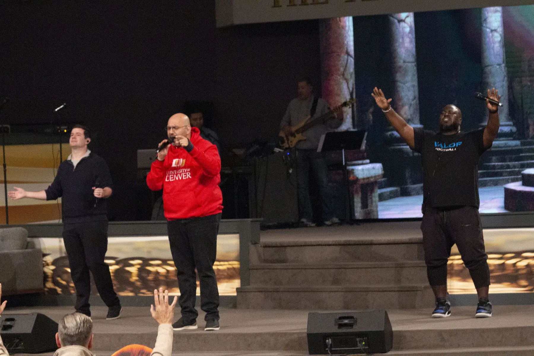 Three men on stage during an event, one wearing a red University of Denver jacket holding a microphone