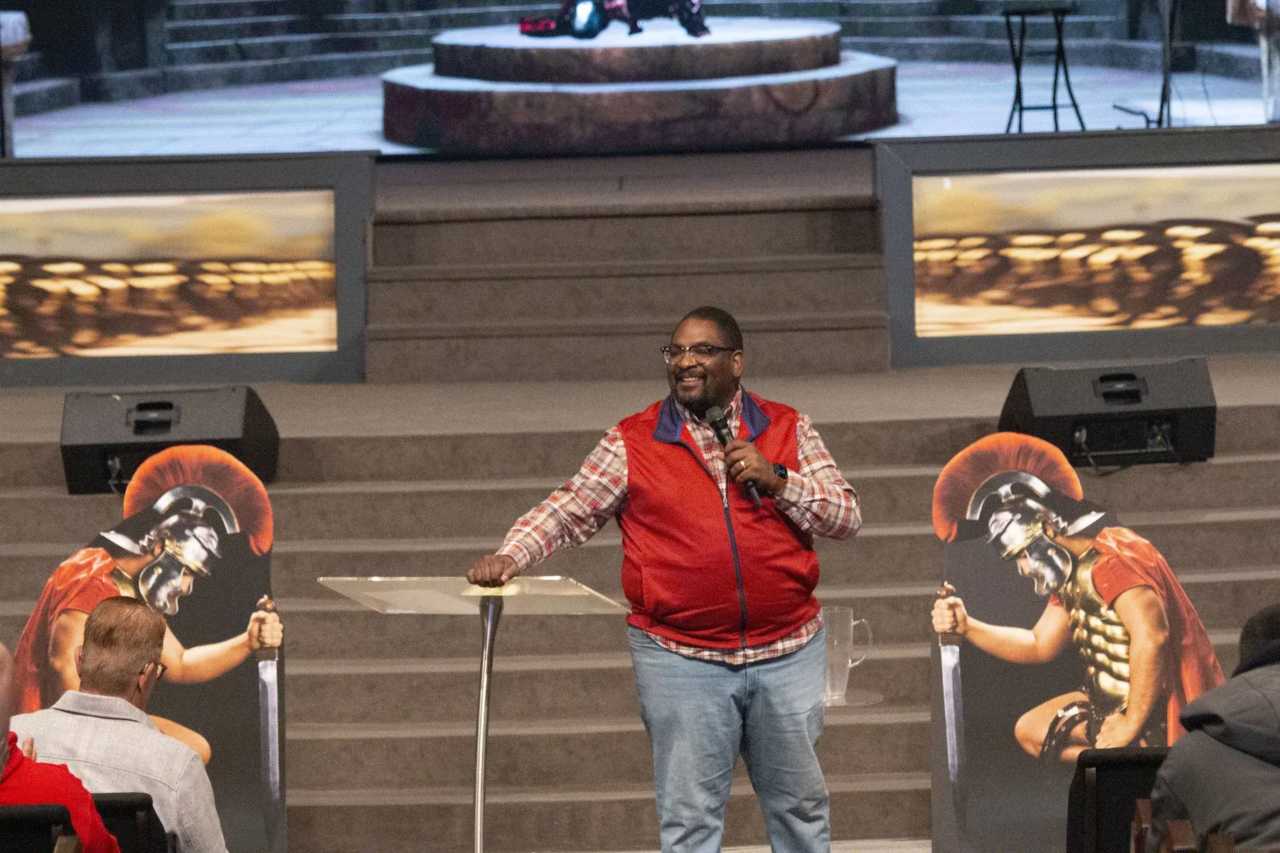 Man speaking at podium on stage with Roman soldier statues flanking him
